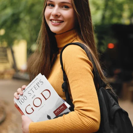 A person holding the devotional book '100 Steps With God' outdoors.